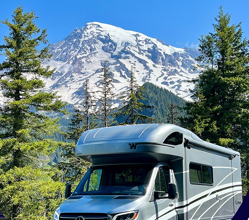 A Winnebago View parked below the snow-covered face of Mount Rainier, framed by tall evergreens.